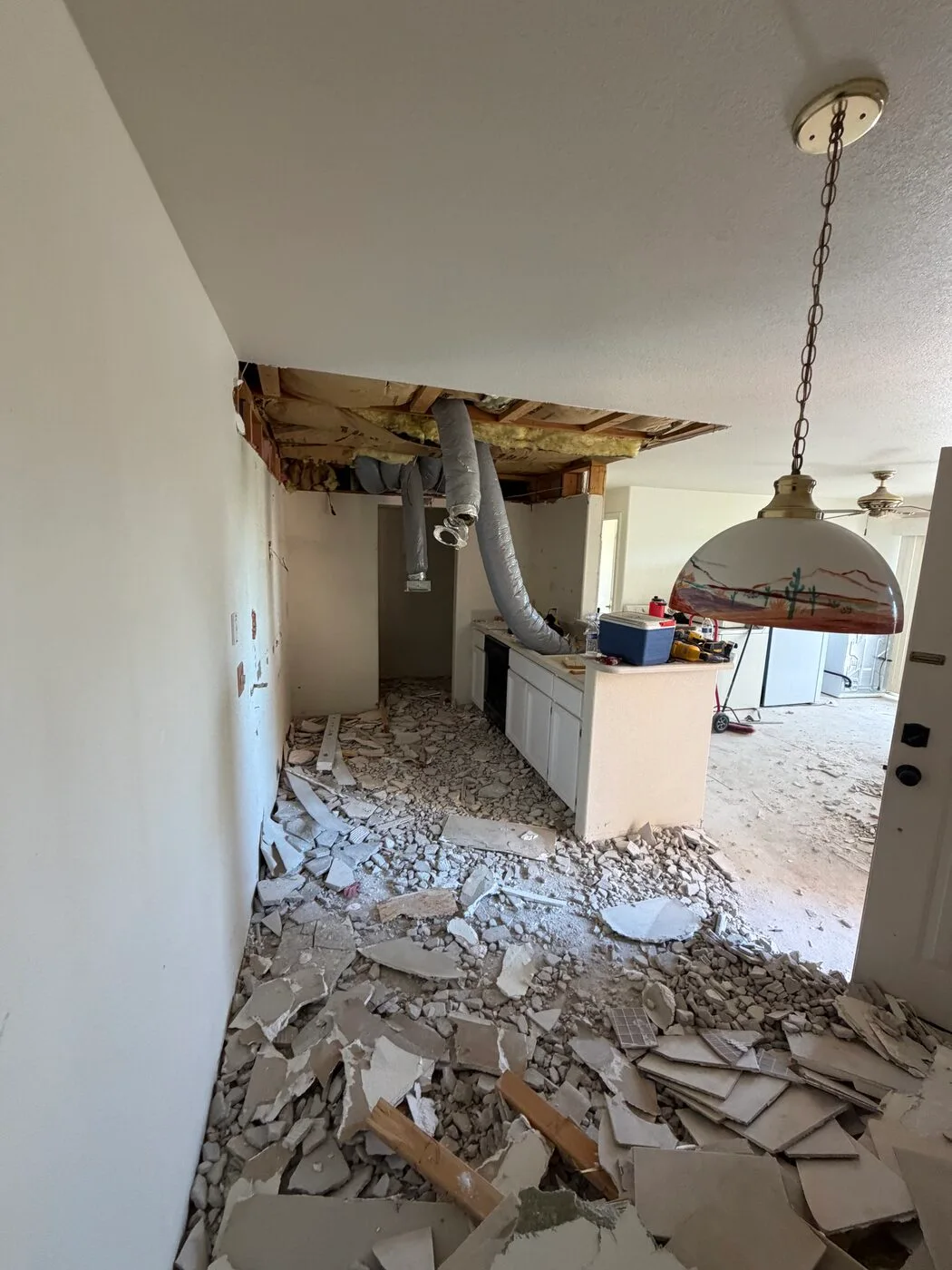Kitchen mid-demolition with soffit opened up, ductwork exposed, and tile and drywall debris covering the floor